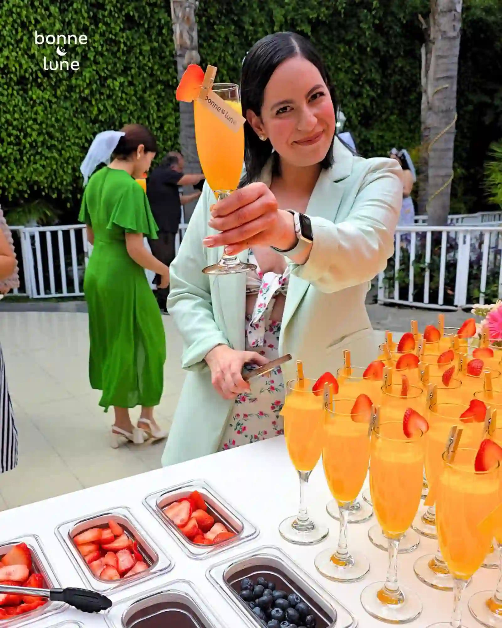 Invitada brindando con una copa de mimosa de Bonne Lune en un evento, barra de mimosas al fondo con más copas servidas